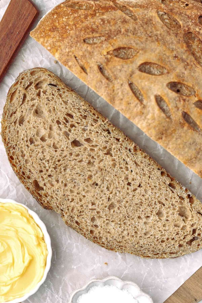 Whole wheat sourdough bread slice with a golden crust, accompanied by butter and salt on a textured surface.